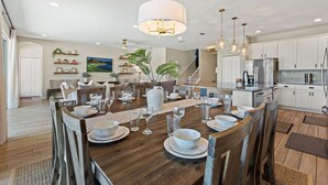 A spacious dining area with a wooden table set for a meal, surrounded by white cabinets and a kitchen in the background.
