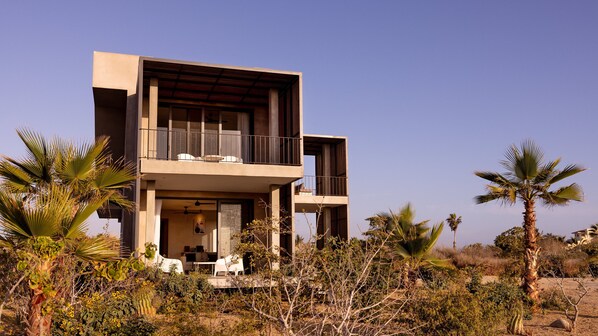 Une maison moderne à deux étages, dotée d’un balcon et entourée de palmiers et de plantes désertiques.