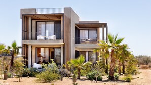 Una casa moderna de dos pisos con un balcón, ventanas amplias y un jardín desértico.