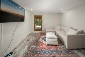A living room with a large flat-screen TV, a beige sectional sofa, a patterned rug, and a wooden floor.