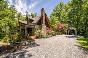 A house with a stone chimney, a covered porch, and a gravel driveway leading to a wooden deck. - Private Log Cabin Near Blowing Rock | Hot Tub, Fire Pit & Creekside Trails (Blowing Rock)