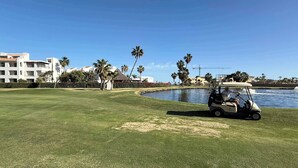 A golf course with a water body, palm trees, and buildings in the background. - Peninsula Luxury Condo Vidanta Golf & Beach Access (Los Cabos)