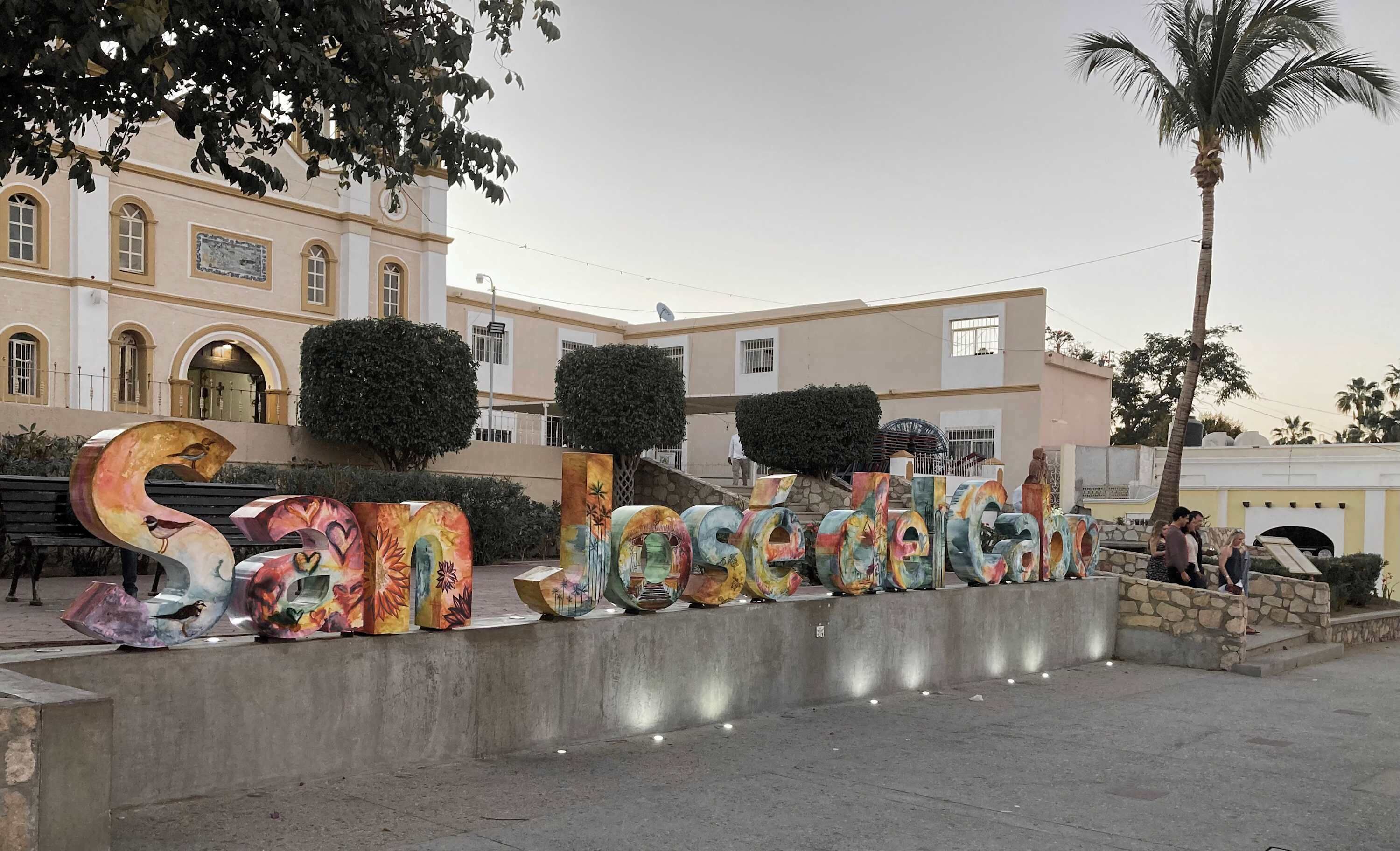 Une enseigne colorée portant le nom « San Jose » en grandes lettres, placée sur le fond d’un bâtiment aux fenêtres en arc de cercle et agrémentée d’un palmier.