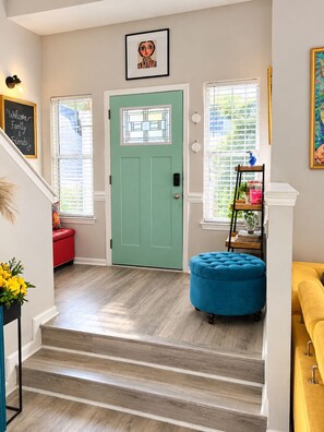 Una escalera conduce a una puerta con ventanal de vidrio esmerilado, un banco rojo, un reposapiés azul y un sofá amarillo.