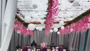 Un espacio de estar al aire libre rústico con bancos de madera y sillas amarillas, decorado con flores rosas colgantes y faroles blancos.
