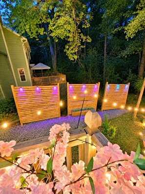 Terraza de madera con luces colgantes, rodeada de follaje y una casa.
