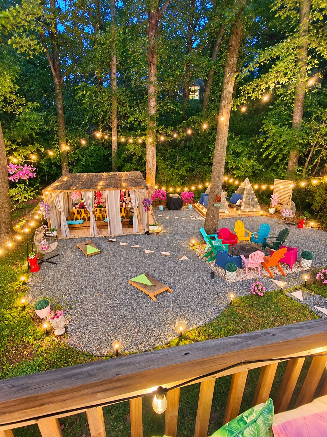 Uma área externa com mesas e cadeiras, deck de madeira, luzes de festa e cadeiras coloridas.