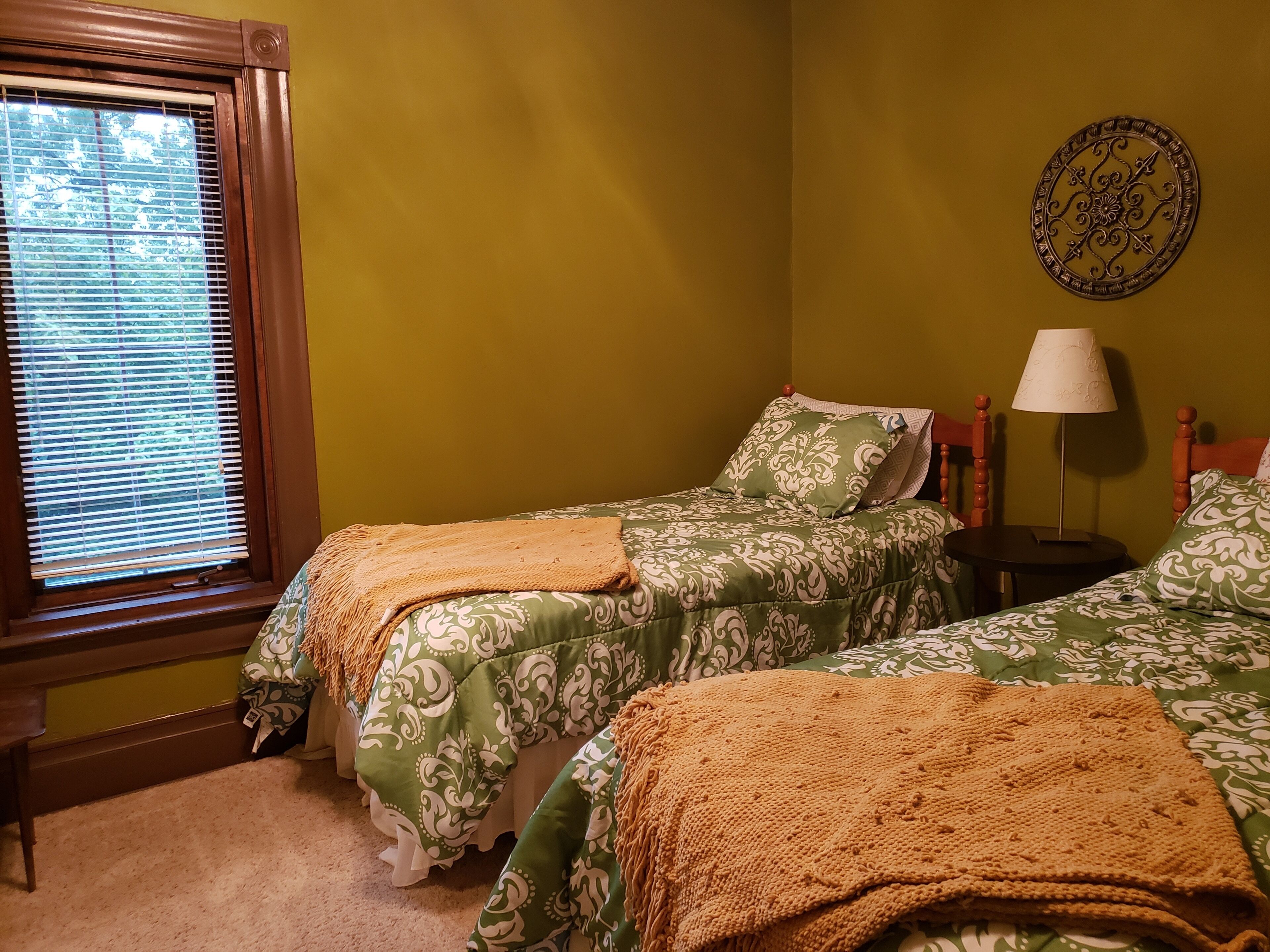 A room with two beds, a window with blinds, a lamp, and a wall decoration.