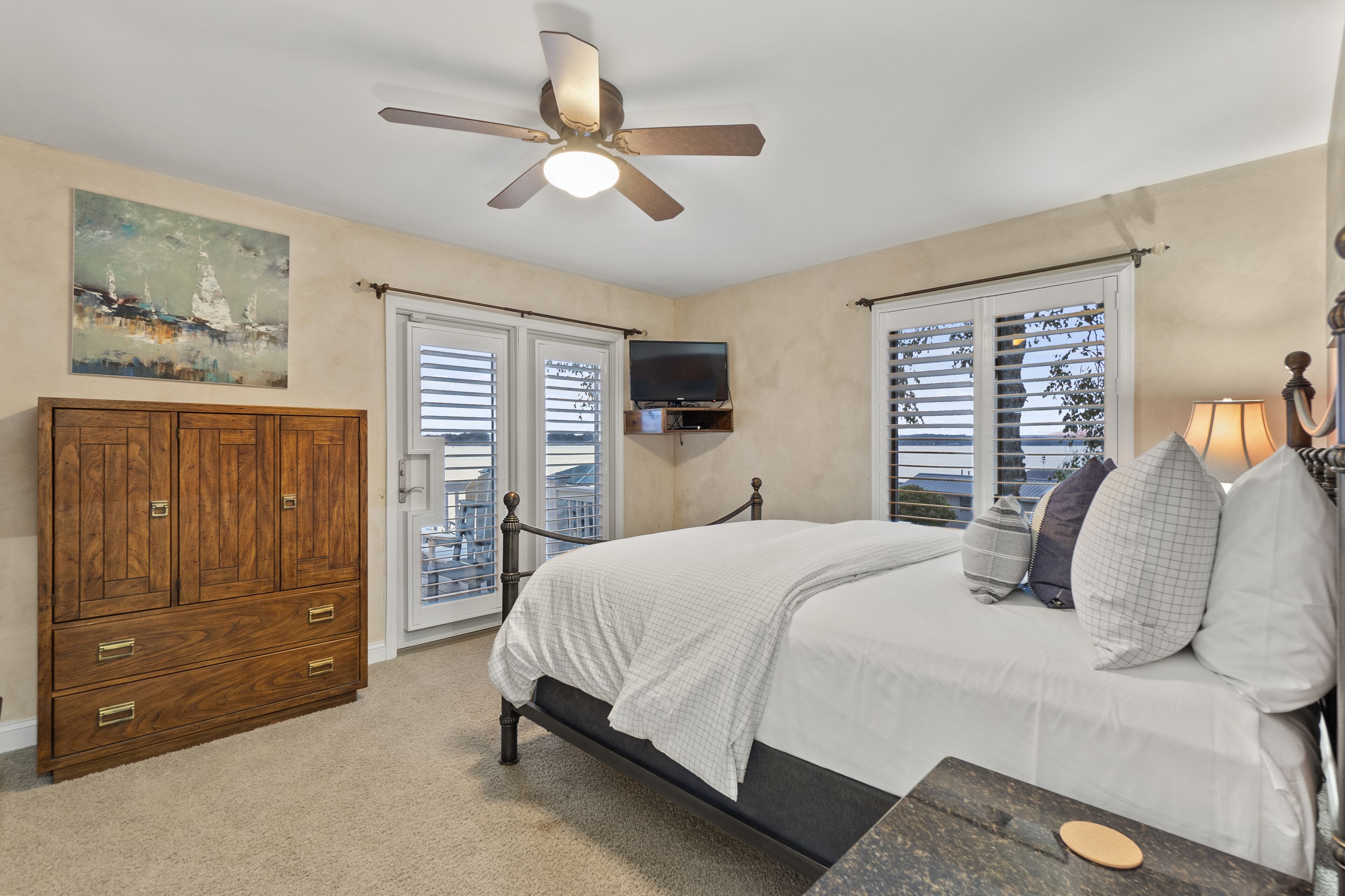 A bedroom with a bed, a wooden wardrobe, a ceiling fan, a TV mounted on the wall, and a window with blinds.