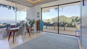 Una sala de estar moderna con un gran ventanal corredero, un escritorio de madera, una silla, un aparador azul y vistas a un paisaje montañoso.
