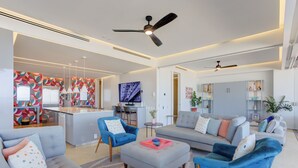 Una sala moderna con un sofá gris, sillones azules y una mesita de centro. Se ve un televisor, un ventilador de techo y una cocina al fondo.
