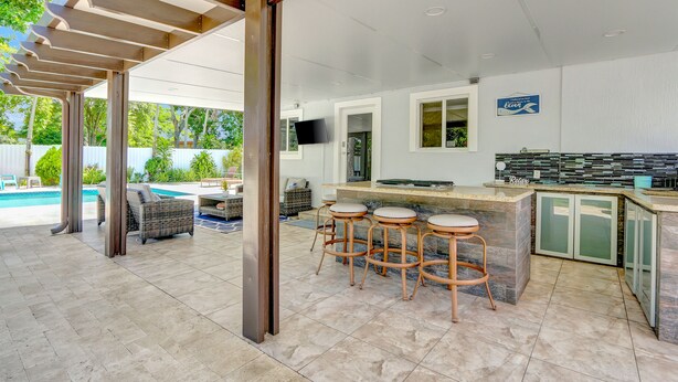 A covered outdoor kitchen area with a pool, bar stools, and a TV. - The Gardens- Private Pool (Miami Gardens)