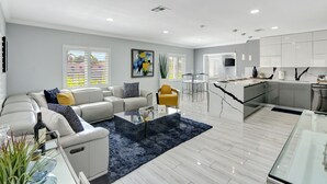 A modern living room with a sofa, coffee table, and a dining area with a kitchen in the background. - The Gardens- Private Pool (Miami Gardens)