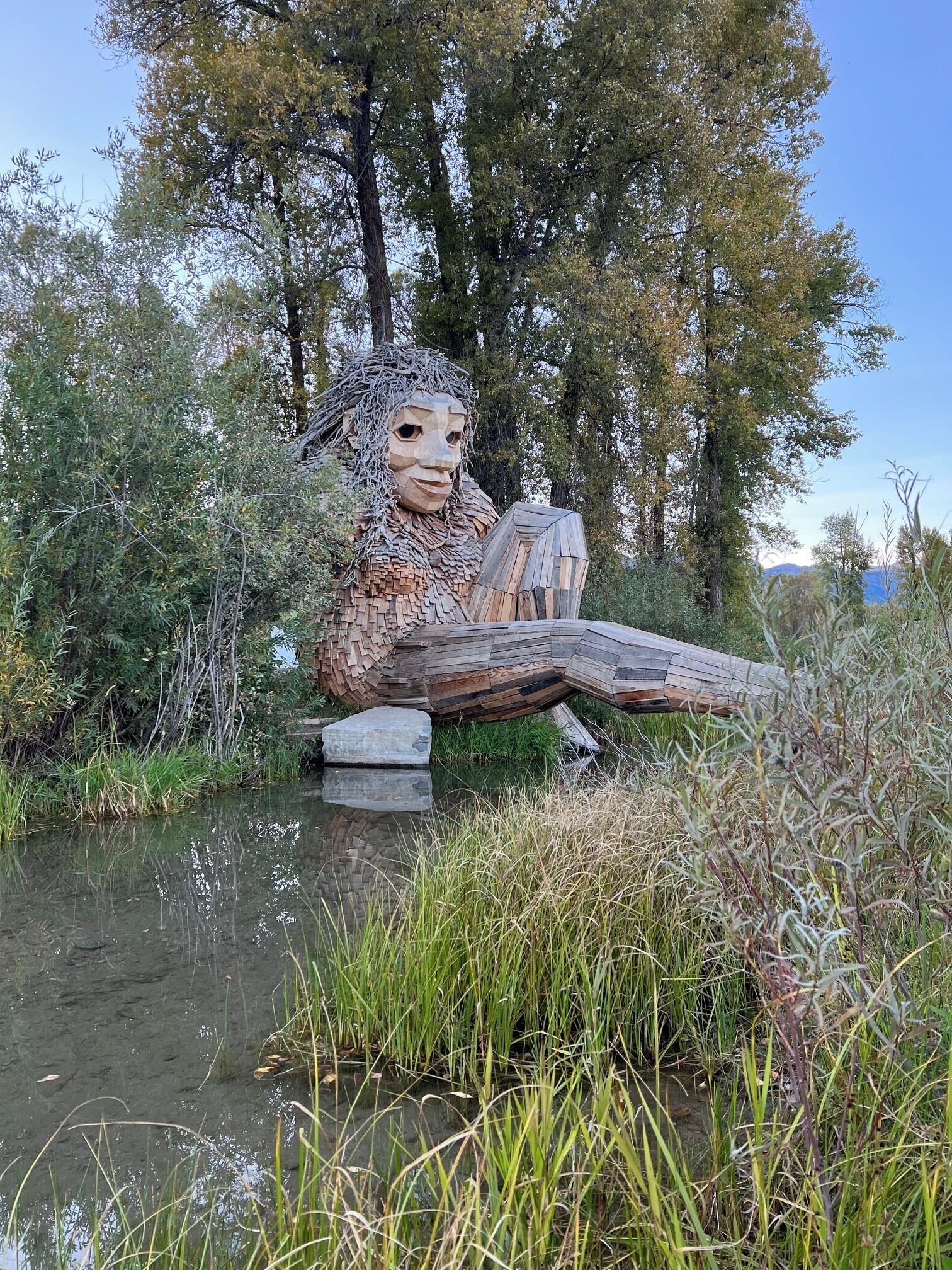 Une sculpture représentant une figure dotée d’une longue queue, assise sur un rocher au bord d’un plan d’eau, entourée d’arbres et de végétation.