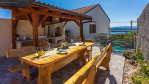 Un'area pranzo esterna in legno con una pergola, una panchina in pietra e vista sul mare.