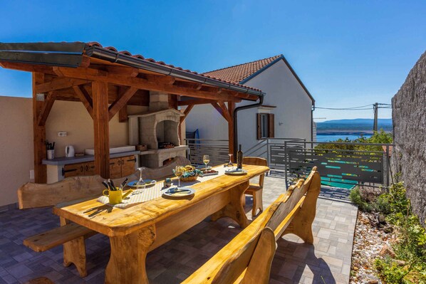 An outdoor wooden dining area with a pergola, a stone bench, and a view of the sea.