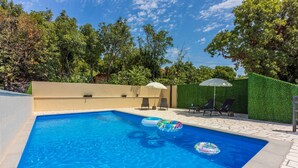 Una piscina rettangolare con acqua cristallina e limpida, circondata da un solarium piastrellato. Si vedono sedie a sdraio e un tavolino con ombrellone.