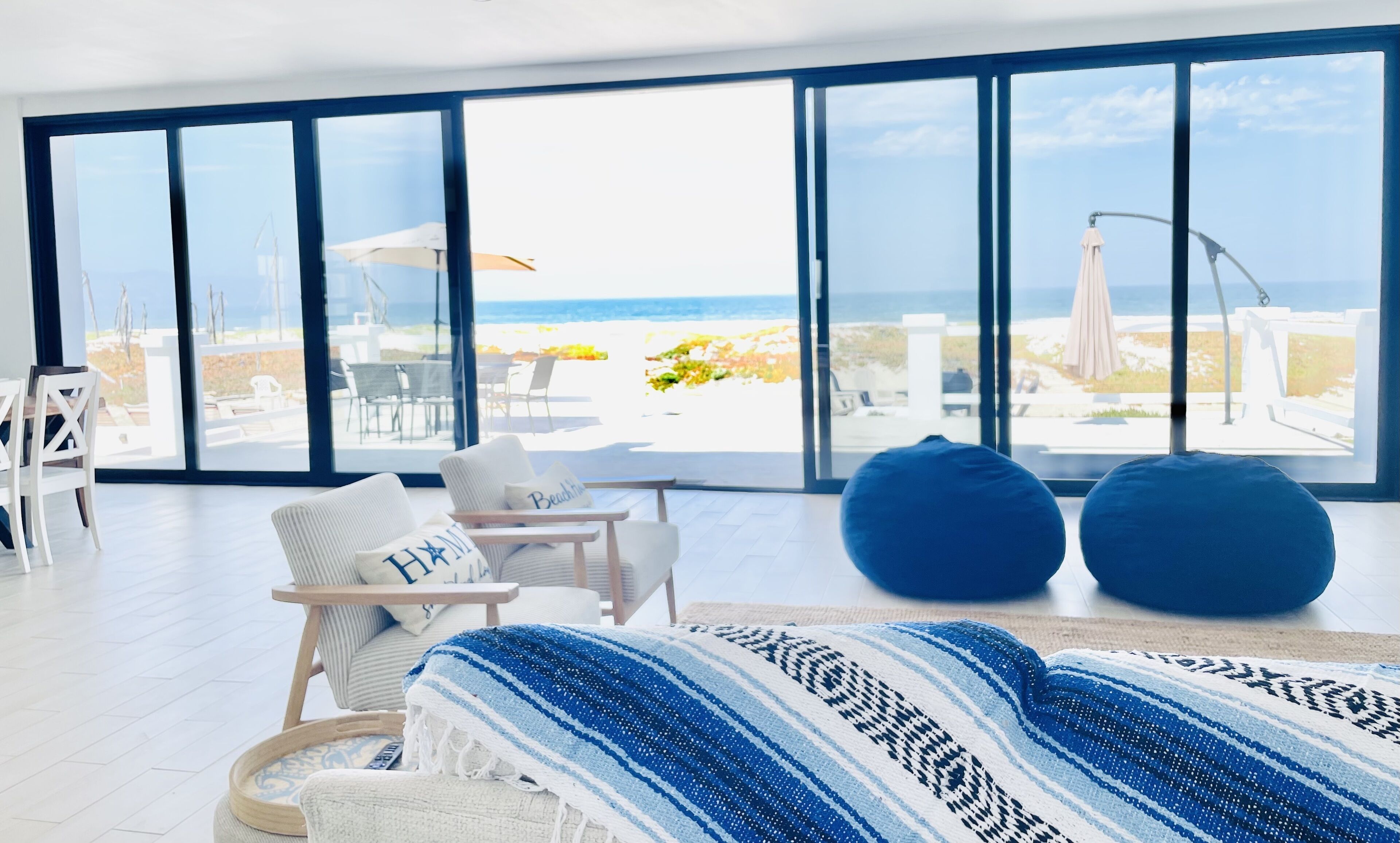 Une pièce spacieuse avec de grandes fenêtres donnant sur une plage, comprenant un lit, des chaises et un coin repas.