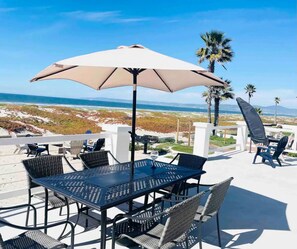 Une terrasse extérieure avec une table et des chaises, un grand parasol et une vue sur la plage et l’océan.