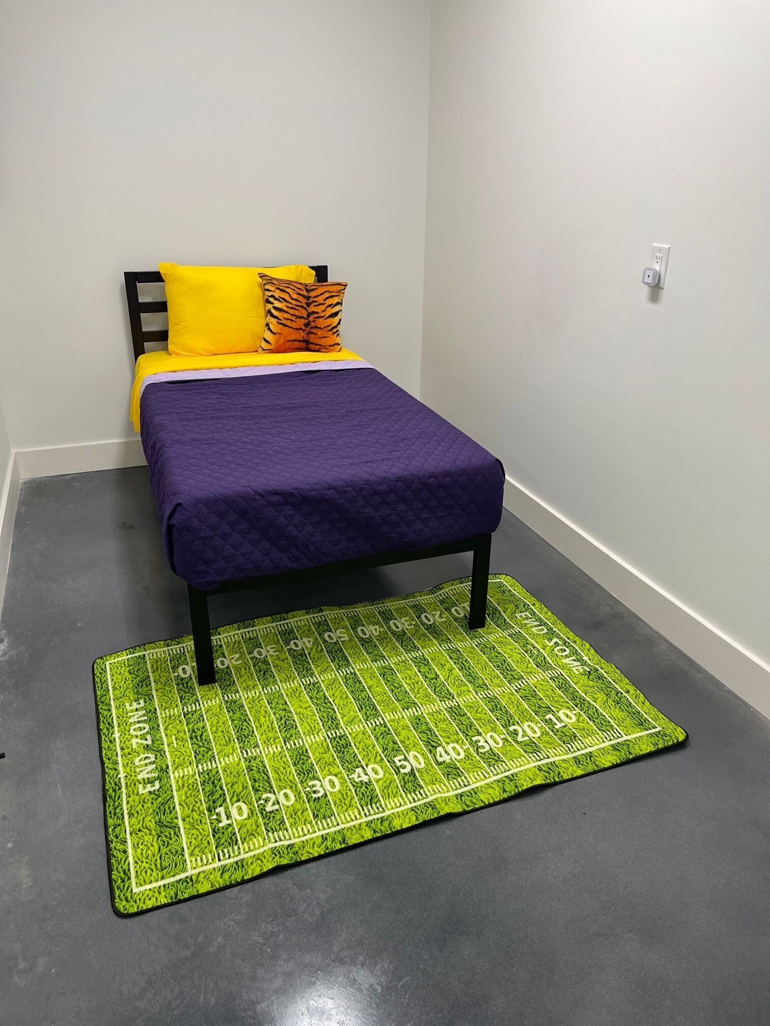 A small room with a bed, a green and yellow rug, and a white wall.