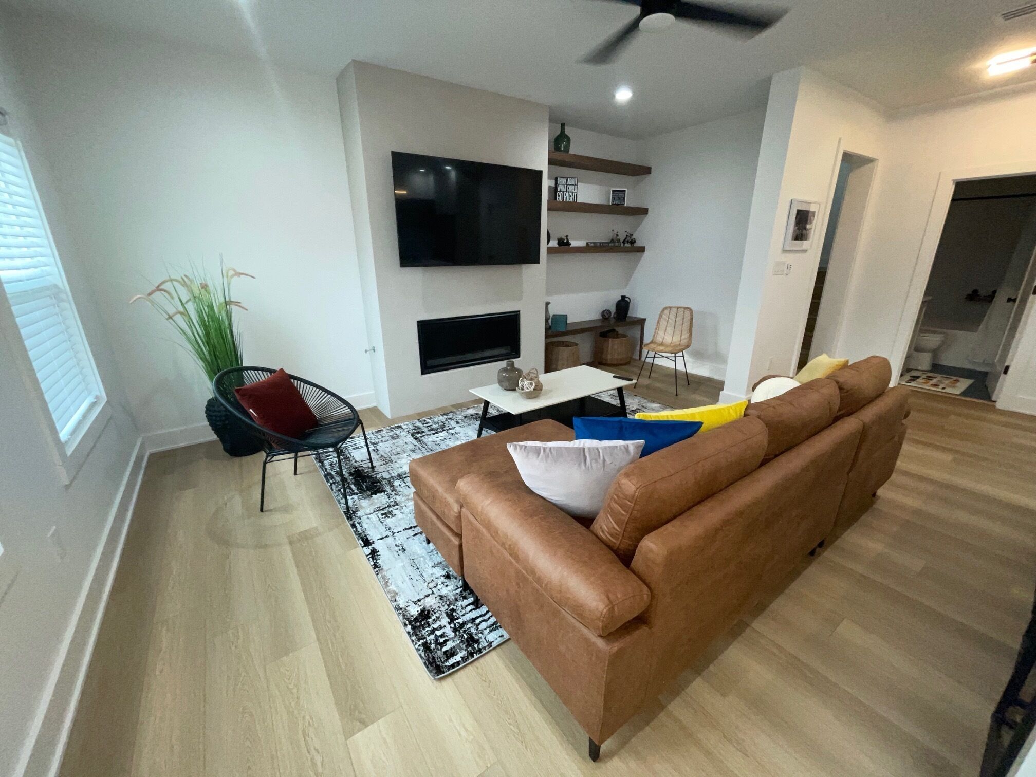 A modern living room with a brown leather sofa, a black and white rug, a dining area with a table and chairs, and a TV mounted on the wall.