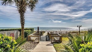 A wooden pathway leading to a beach with a palm tree and a fence.