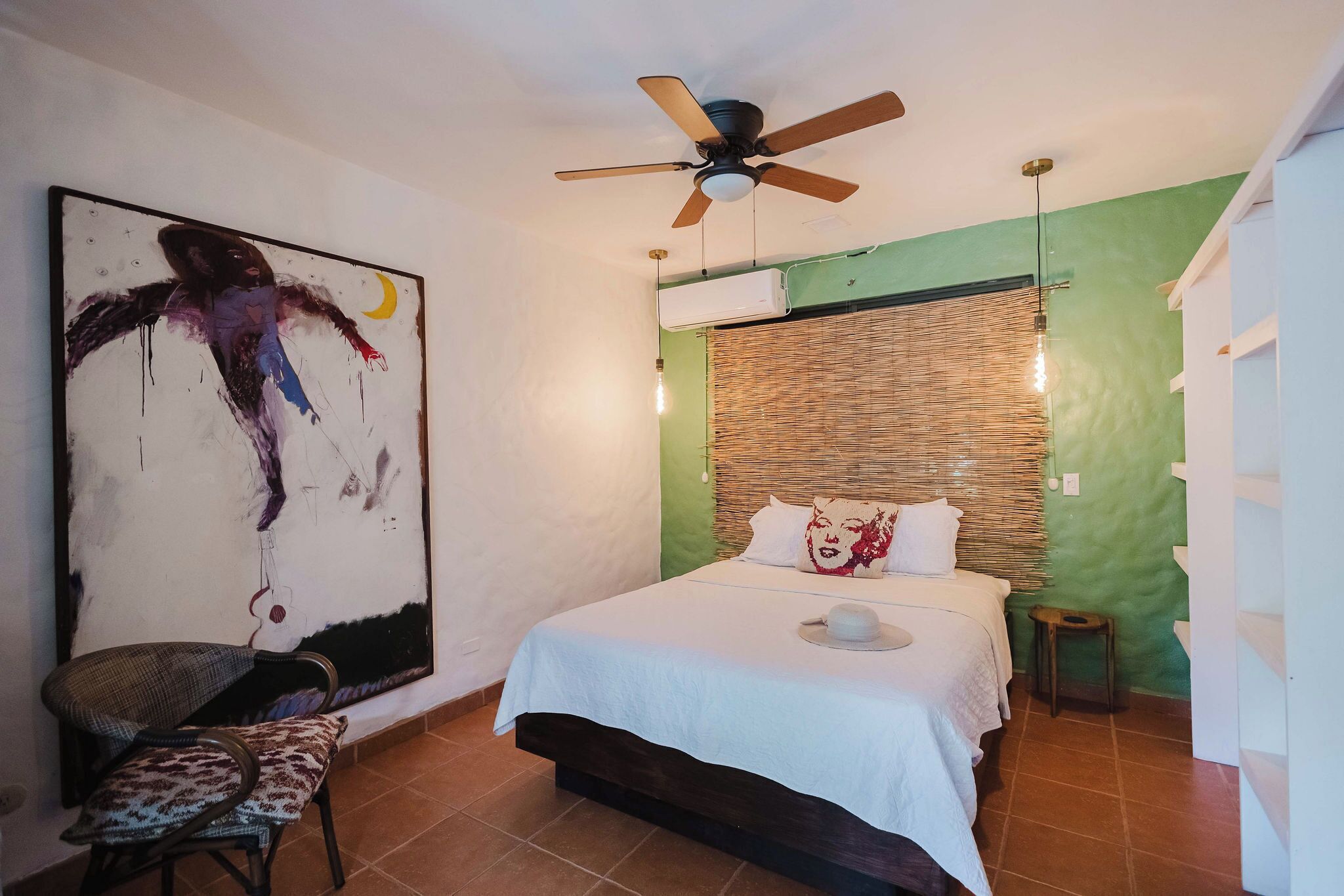 Un dormitorio con cama, ventilador de techo, aire acondicionado en la pared y un cuadro abstracto de gran tamaño.