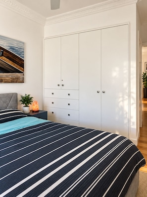 A bedroom with a bed, a nightstand with a plant, a wall-mounted heater, and a wardrobe with multiple doors. - Seaside Studio (Elwood)