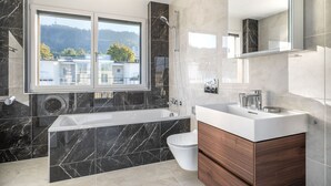 Un baño moderno con bañera, inodoro y lavamanos. Se aprecia una ventana con vistas a edificios y árboles.