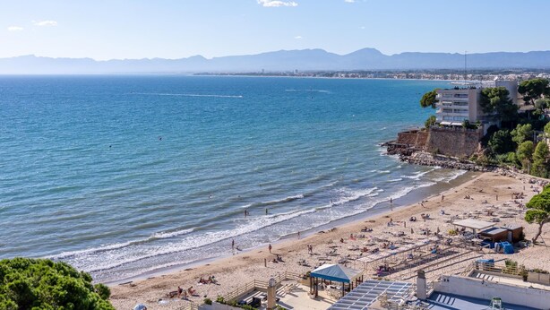 Une plage côtière avec un rivage rocheux, une zone de plage sablonne agrémentée de parasols et de chaises longues, et une vue dégagée sur la mer et les montagnes lointaines.