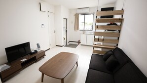 A modern living room with a flat-screen TV, a wooden coffee table, a black sofa, and a kitchen area with a window. - KARCHIBE ISHIGAKI (Ishigaki)