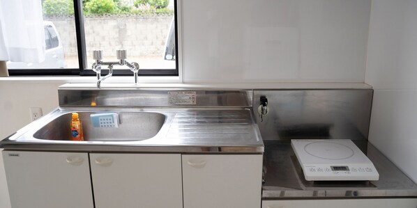 A kitchen with a stainless steel sink, a dish soap bottle, a weighing scale, and a window with a view of a parked car and greenery outside.