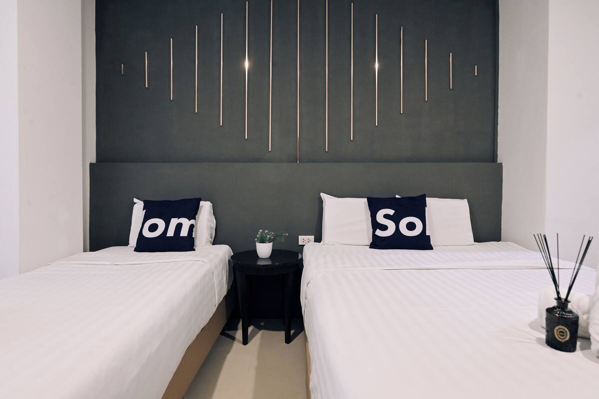 A hotel room with two beds, each with white bedding and pillows with "om" and "so" text. There is a small table with a potted plant and a black vase with reed diffusers.