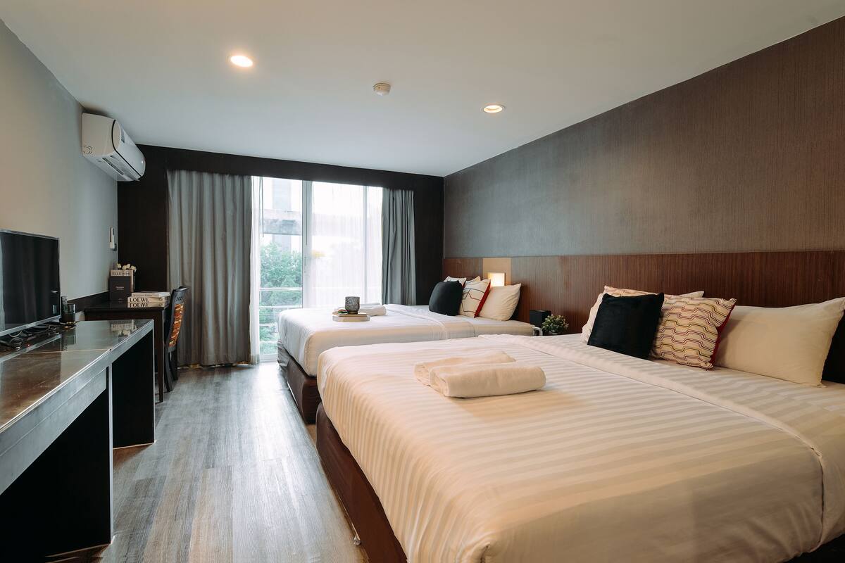 A hotel room with two beds, a desk, a TV, and a large window with curtains.