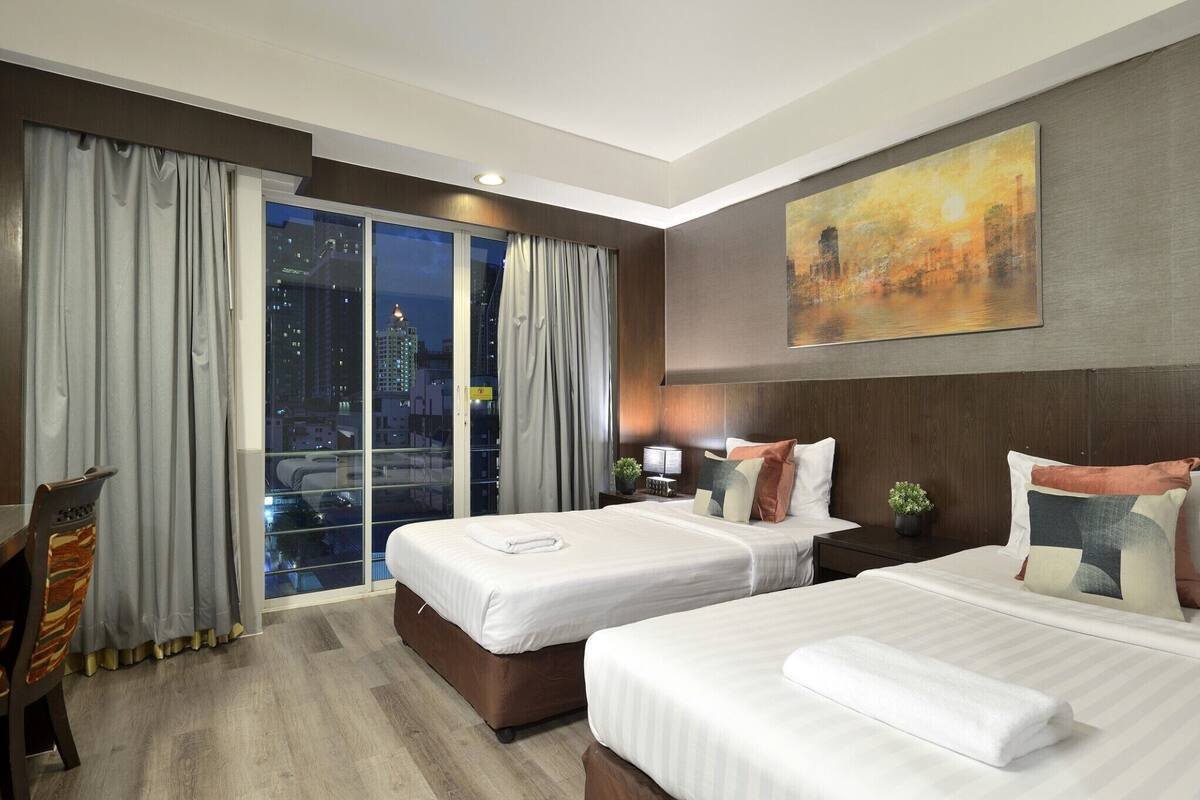 A hotel room with two beds, a city view, a painting on the wall, and a dining area with a chair.