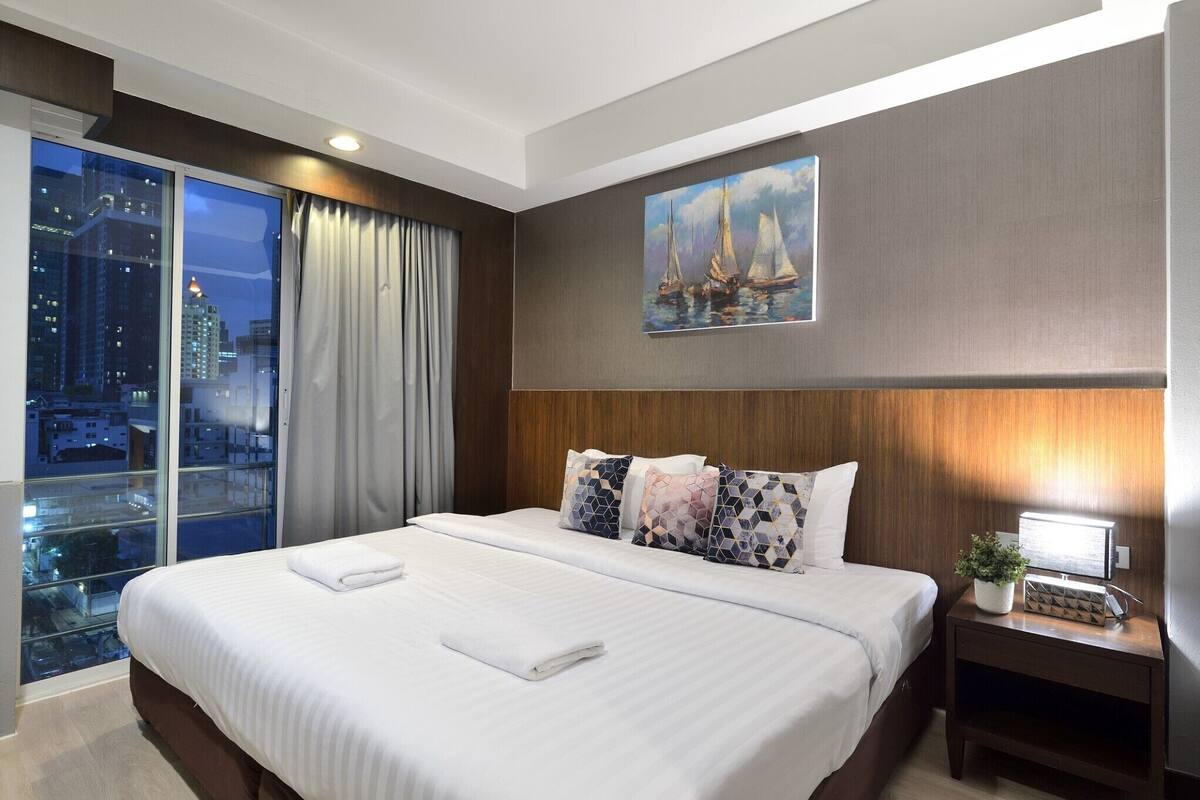 A hotel room with a large bed, a nightstand with a lamp, a painting of sailboats, and a view of the city through large windows.