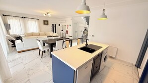 Een moderne keuken met een centraal eiland, een eethoek met een witte tafel en stoelen, en een zithoek met een bank en gordijnen.