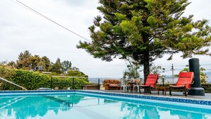 Una piscina con borde azul, rodeada de sillas reclinables rojas y un árbol grande.
