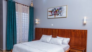 A bedroom with a wooden headboard, a bed with white linens, a nightstand with drawers, teal curtains, and a wall-mounted light fixture. - Beautiful 1-Bed apartment in the heart of Kampala (Kampala)