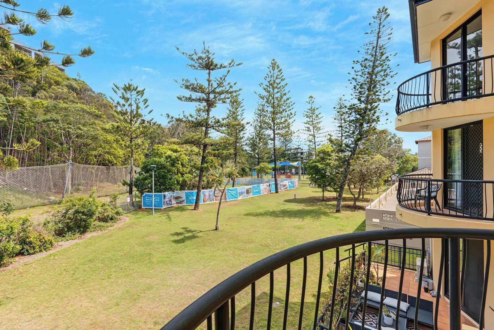 A balcony with a view of a landscaped area, including trees and a fenced playground.