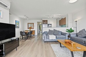 A modern living room with a flat-screen TV, grey sofas, and a dining area with a wooden table and chairs.