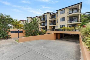 A multi-story residential building with balconies, a parking area, and surrounding greenery.