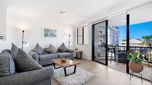 Een moderne woonkamer met een grijze bank, gekleurde kussens, een houten salontafel en een wit vloerkleed. Een groot raam met uitzicht op gebouwen en een balkon met tuinmeubilair is zichtbaar.