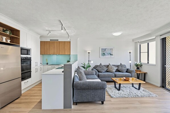 A modern kitchen with white cabinets, a grey sofa, and a wooden dining table.
