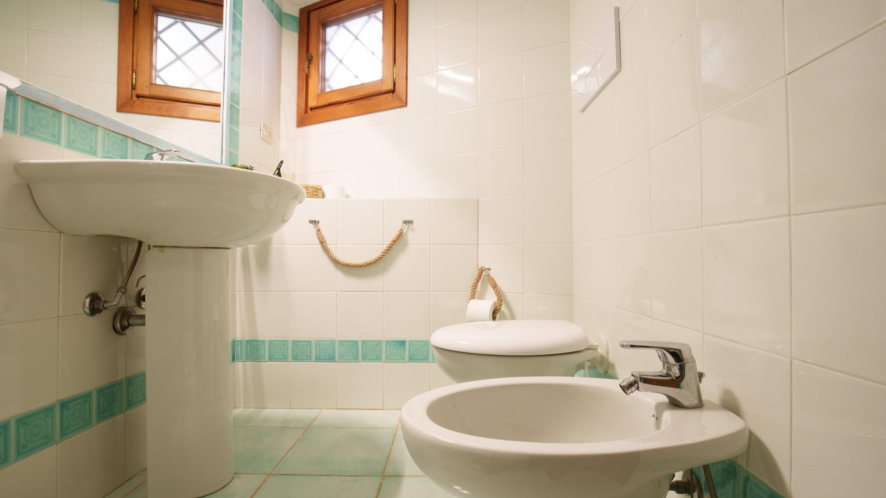 Un baño con un lavamanos blanco, un inodoro y un bidé. Hay dos ventanas con marcos de madera.