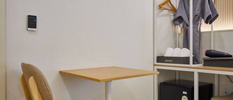 Habitación pequeña con una silla de madera, una mesita, un armario con ropa colgada y un electrodoméstico negro.
