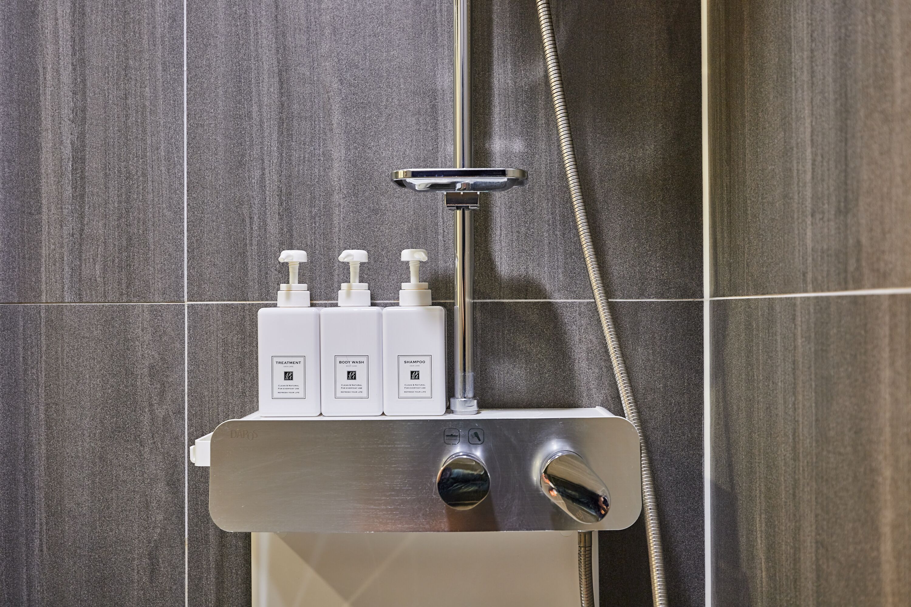 A shower area with a handheld showerhead, a control panel, and three bottles of toiletries.