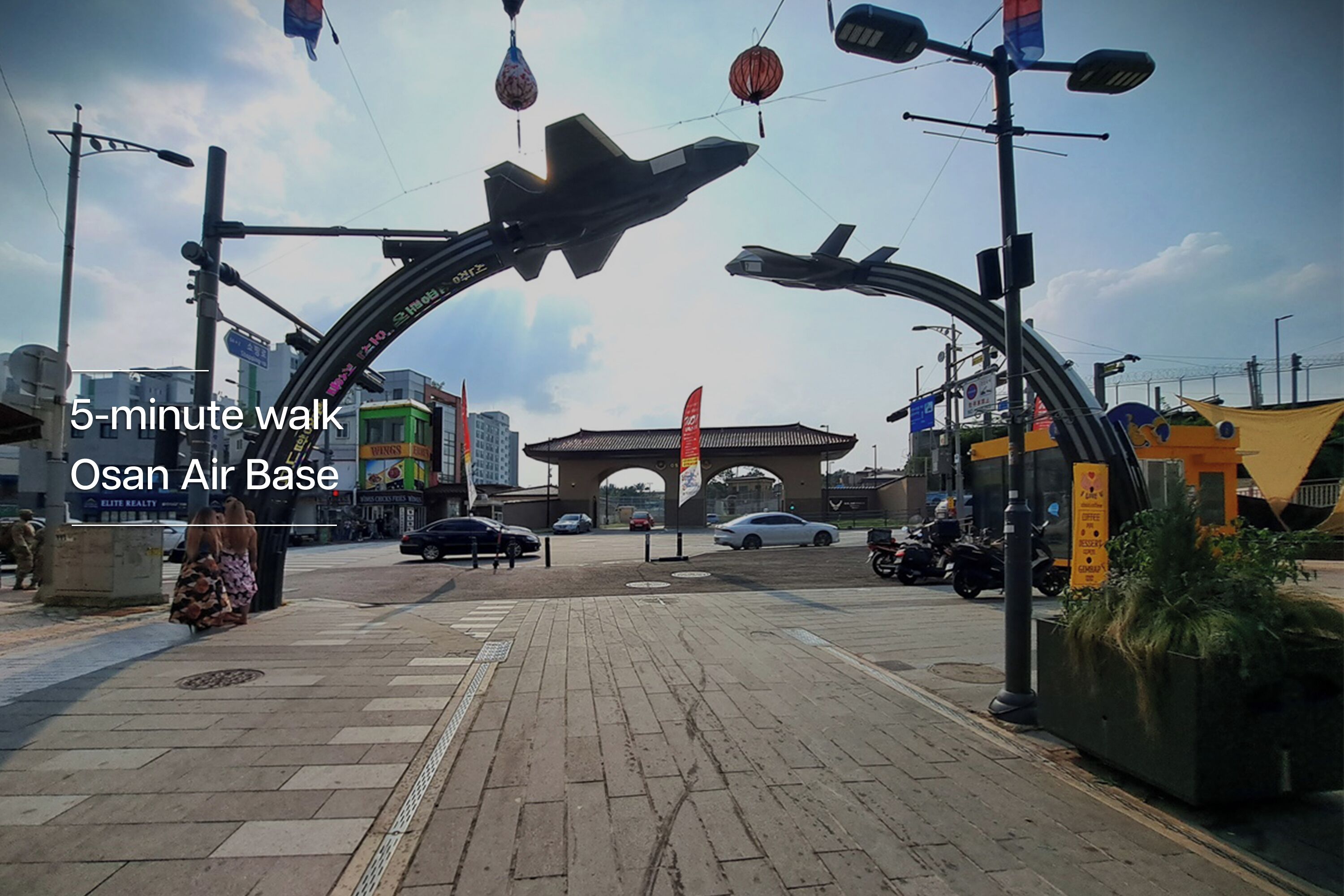A paved walkway with a large archway, a jet, and a banner reading "5-minute walk Osan Air Base."