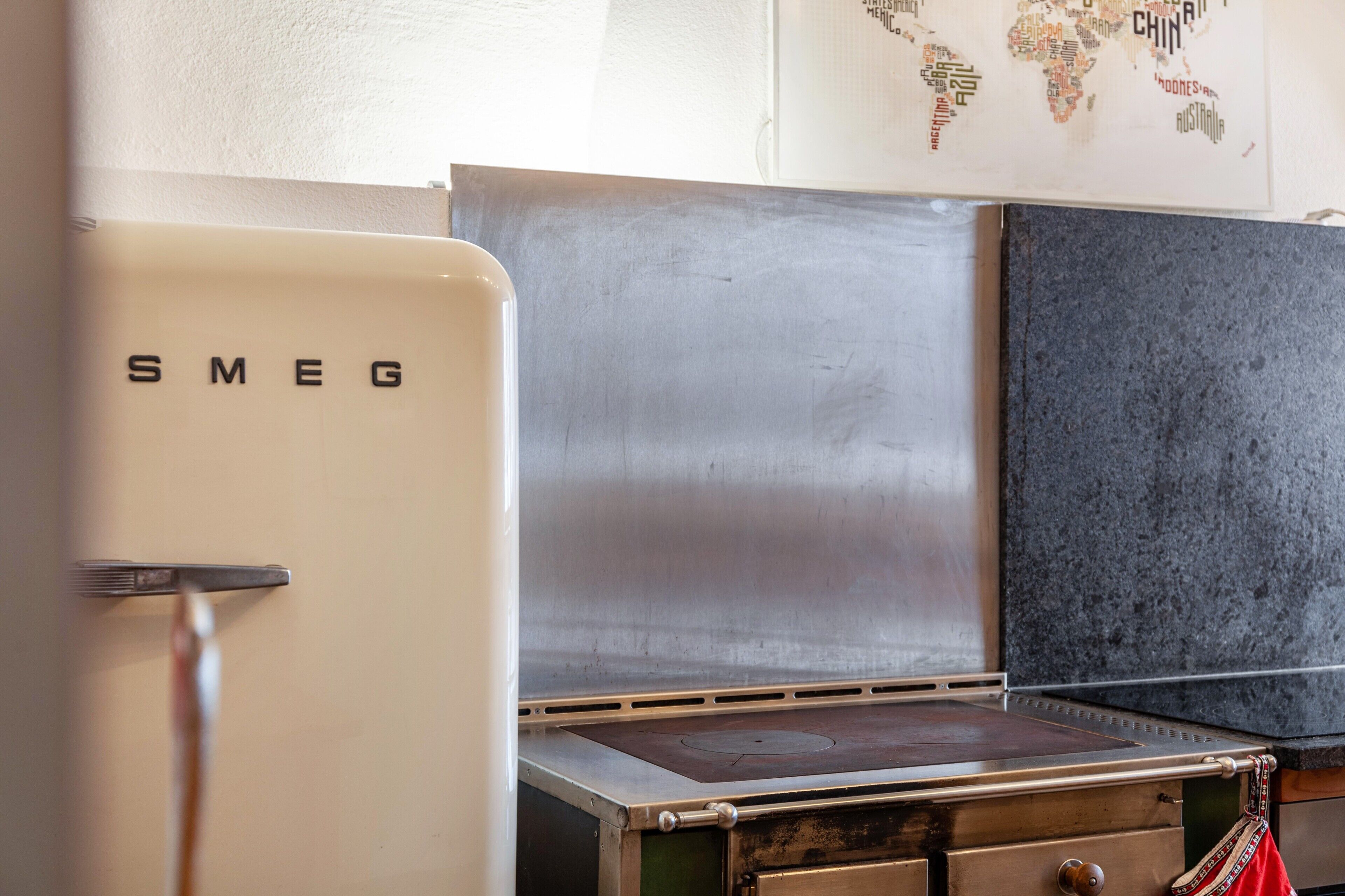 A kitchen with a stainless steel range hood, a white refrigerator with 'SMEG' on it, and a world map on the wall.