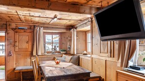 Interno di una baita in legno con un tavolo da pranzo, sedie, una televisione e una finestra con tende.